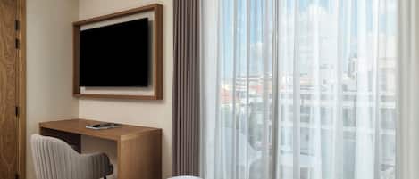 A modern hotel room with a wooden desk, a grey chair, a small ottoman, a flat-screen TV mounted on the wall, and a large window with sheer curtains.