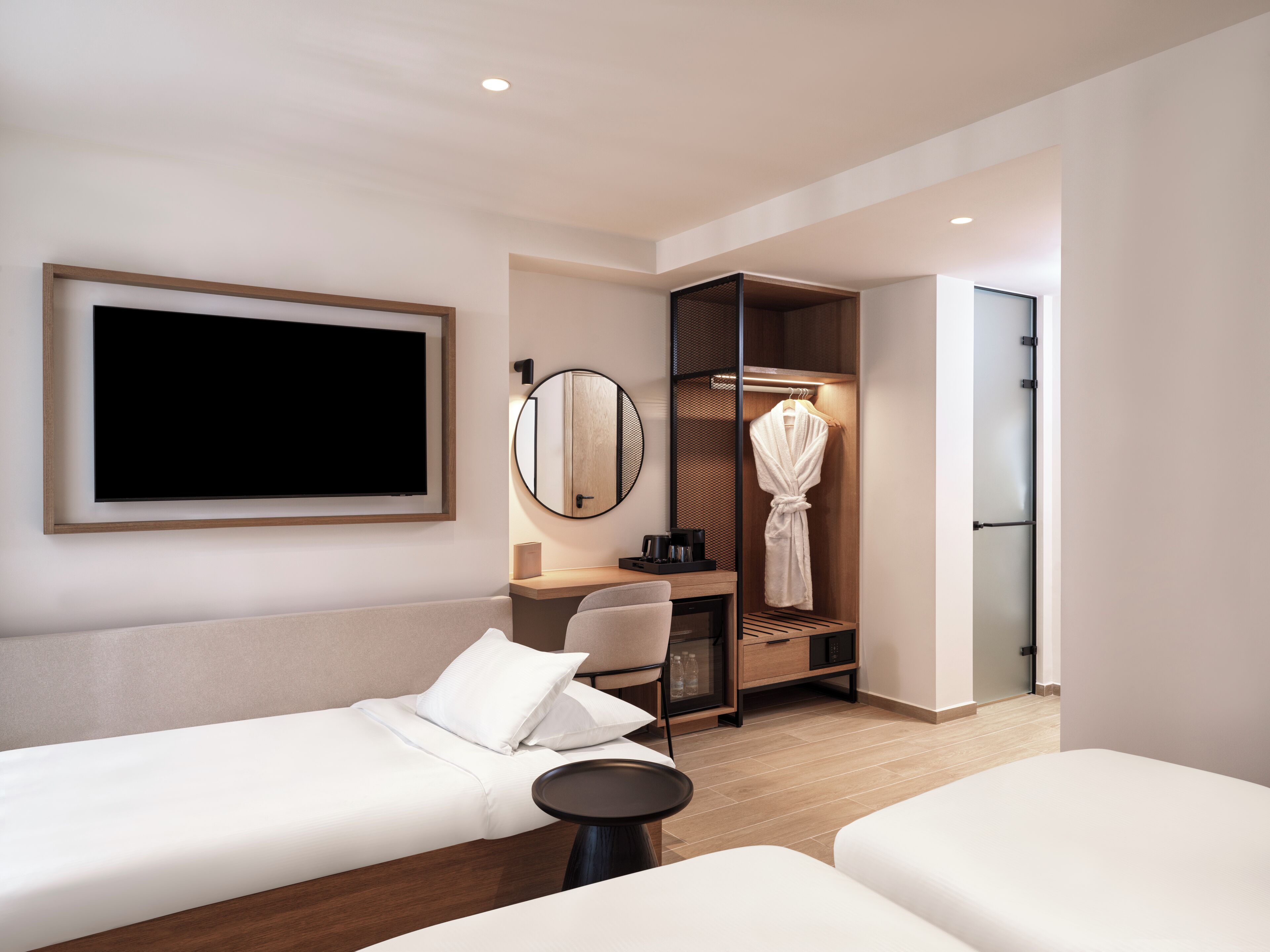 Une chambre d’hôtel moderne avec deux lits simples, un bureau avec une chaise, un peignoir suspendu à un porte-manteau et un grand miroir.