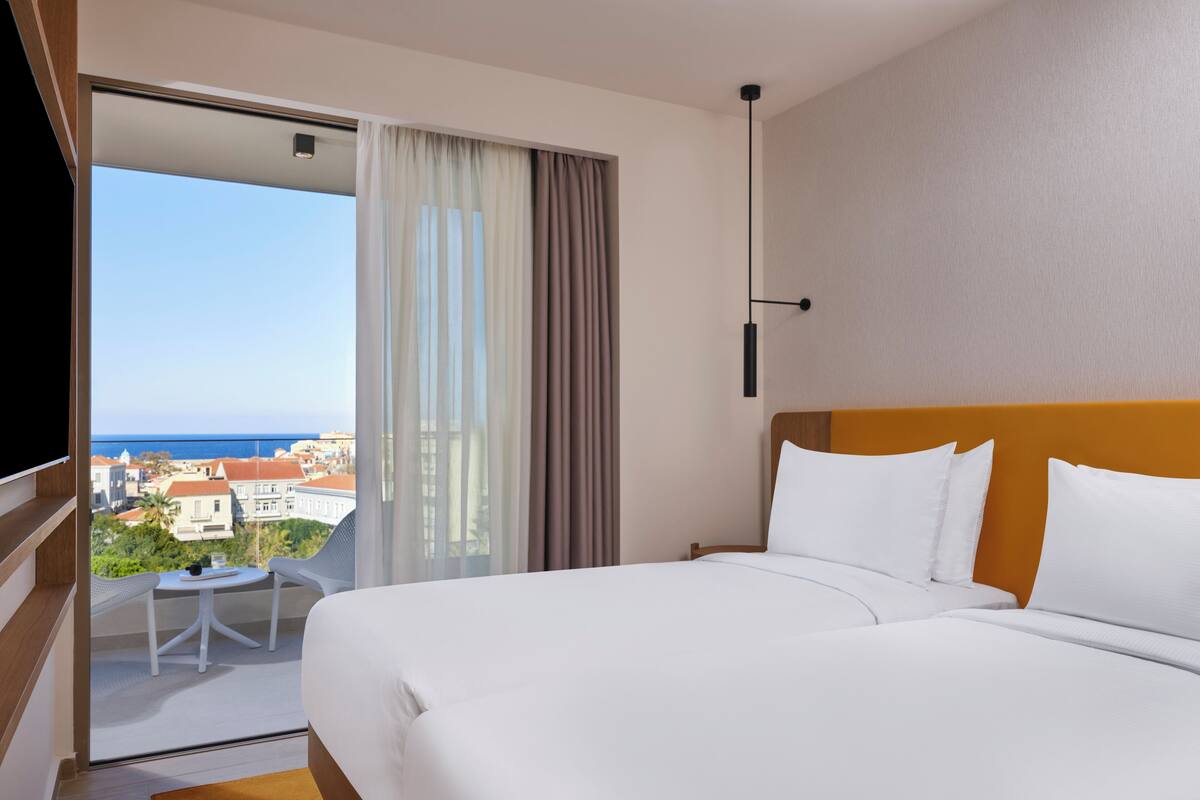 A hotel room with a bed, a small table, and a view of the city and sea.