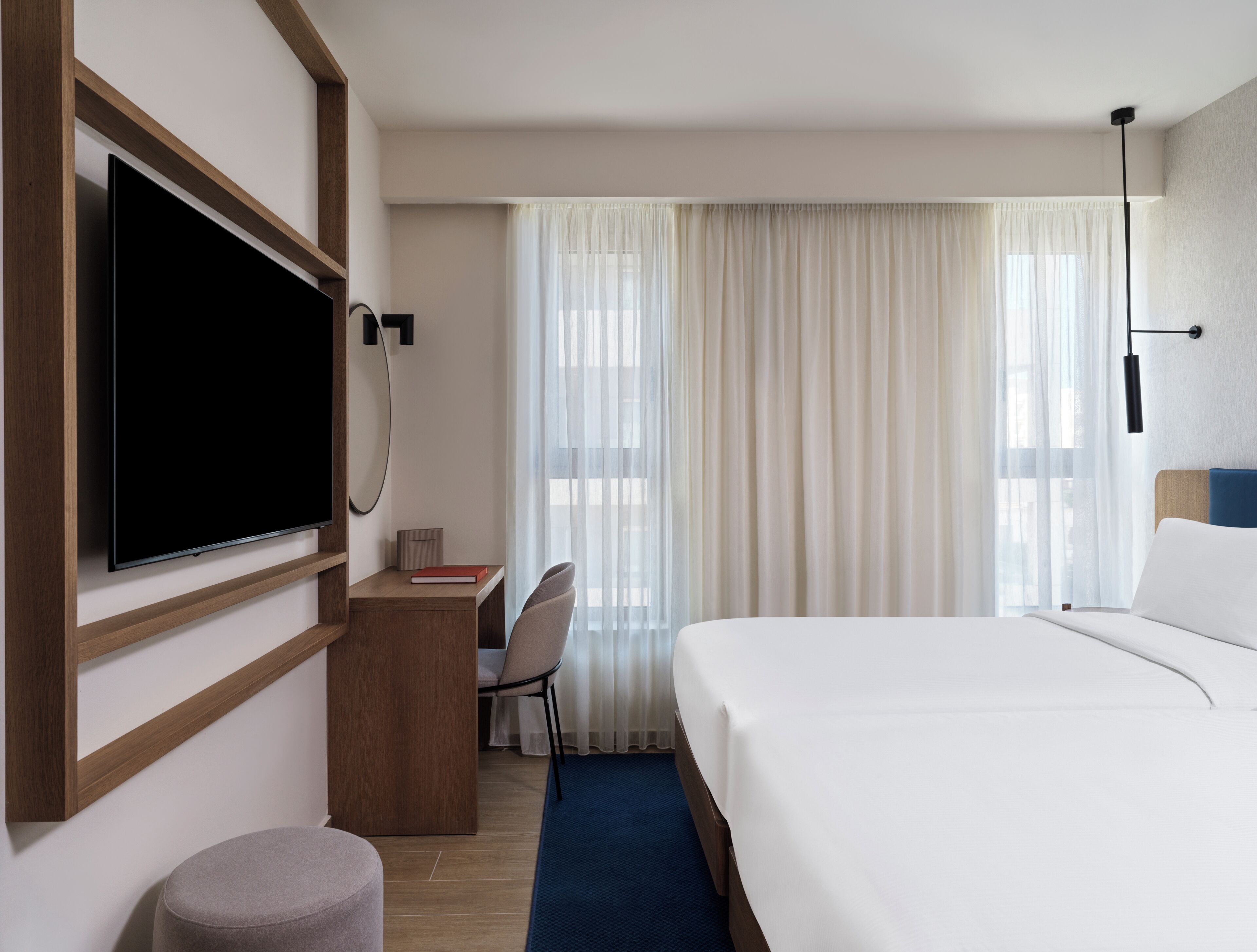 A hotel room with a bed, a desk with a chair, a television, and a window with curtains.
