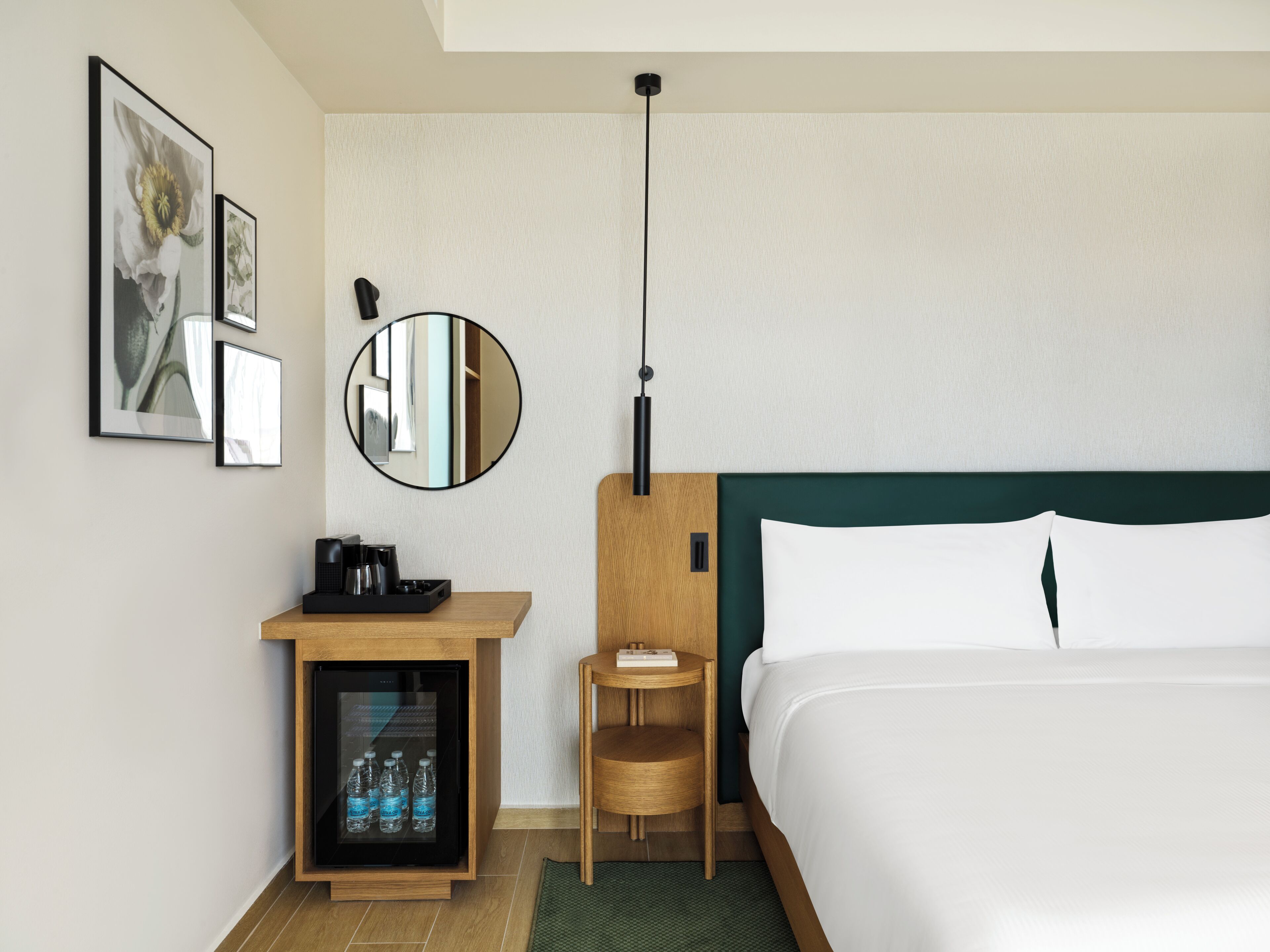 A hotel room with a bed, a small wooden desk with a coffee maker, a refrigerator with water bottles, a round mirror, and framed pictures on the wall.