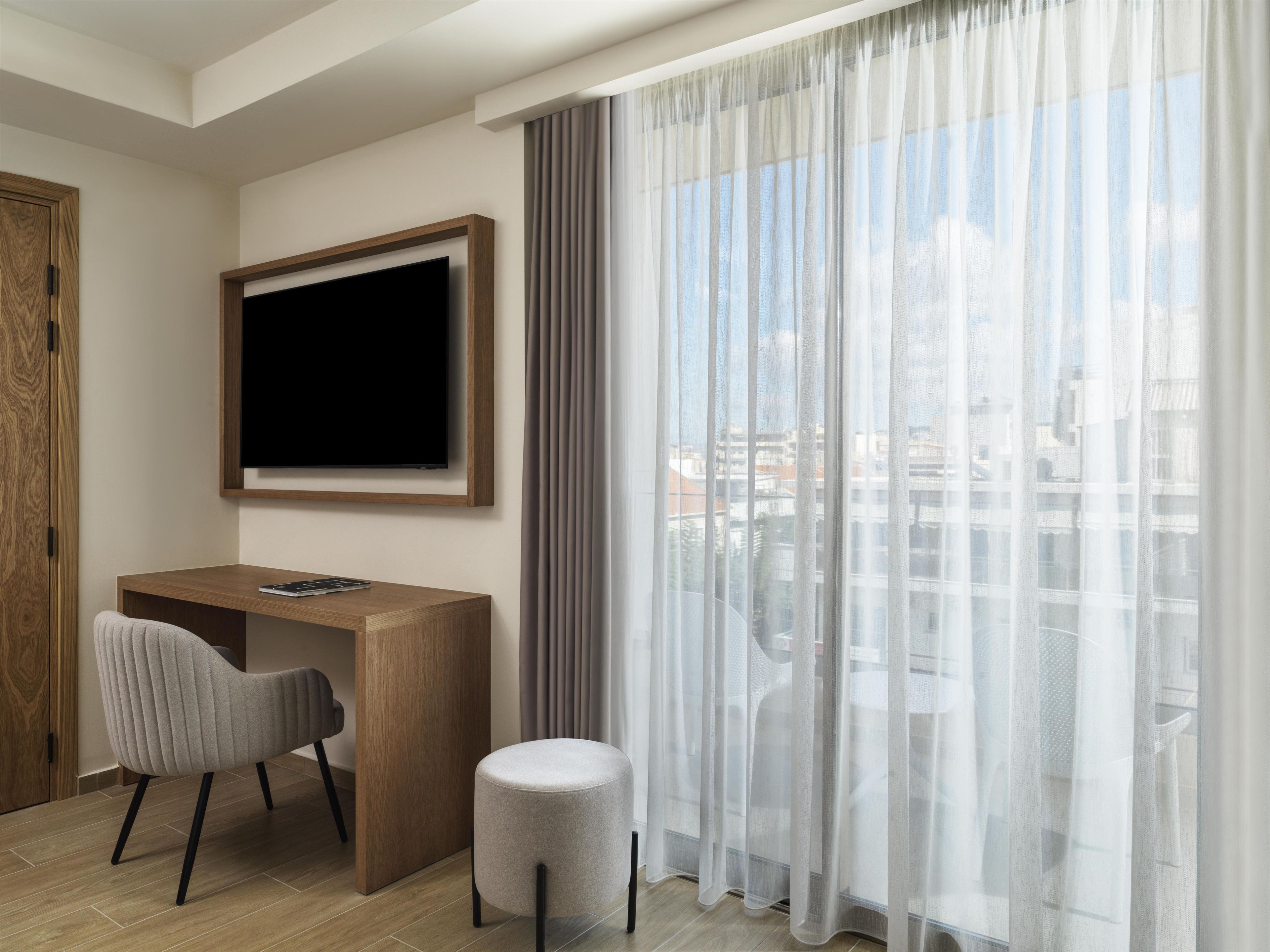 Quarto de hotel com escrivaninha de madeira, cadeira, televisão presa na parede e uma grande janela com cortinas finas.