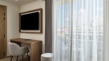 Quarto de hotel com escrivaninha de madeira, cadeira, televisão presa na parede e uma grande janela com cortinas finas.