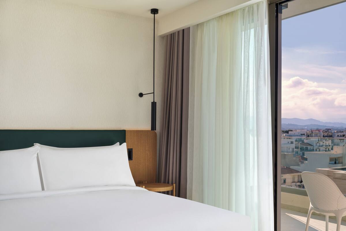 A hotel room with a large bed, a view of the city through a window, and a balcony with a chair.