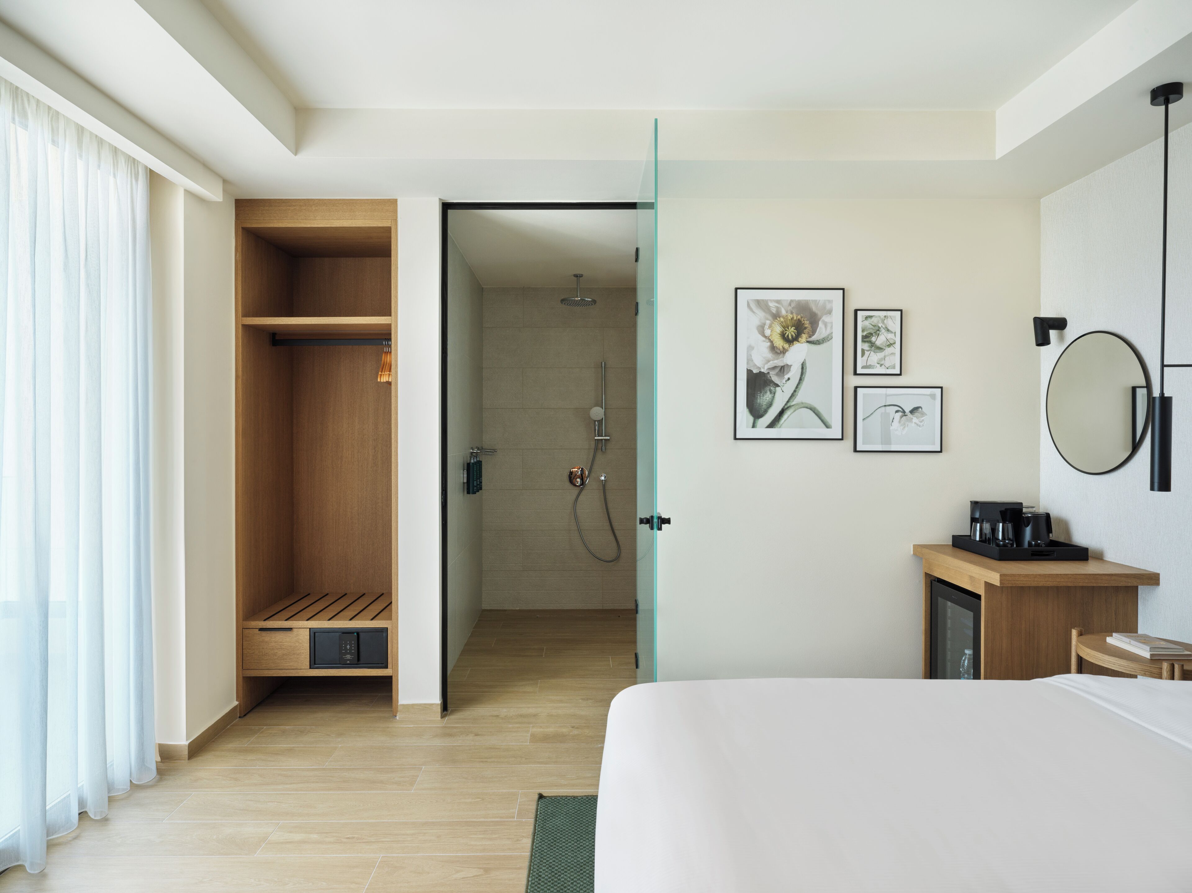 A modern hotel room with a wooden wardrobe, a shower area, a bed, a bedside table with a coffee maker, and framed artwork on the wall.
