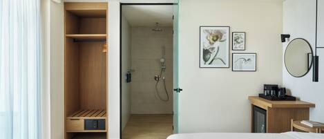 A modern hotel room with a wooden wardrobe, a shower area, a bed, a bedside table with a coffee maker, and framed artwork on the wall.