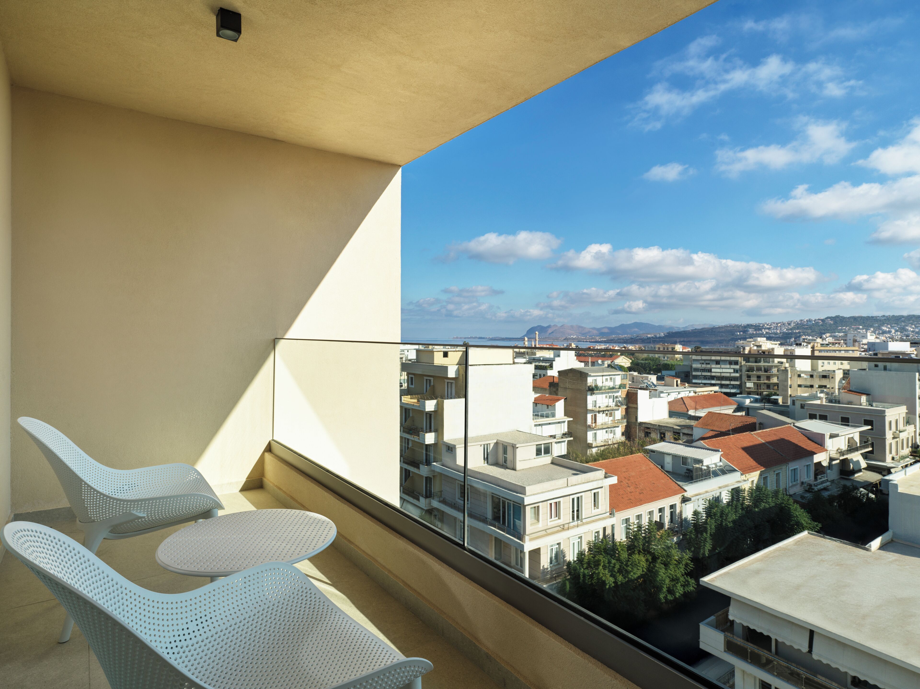 Ein Balkon mit zwei weißen Stühlen und einem Tisch, mit Blick auf eine Stadtlandschaft mit Gebäuden und blauem Himmel.