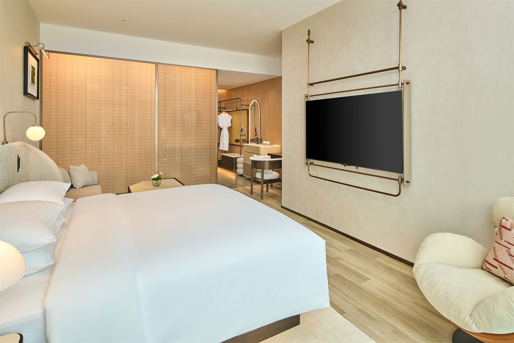 Una habitación de hotel moderna con una cama grande, un televisor montado en la pared, un baño independiente con tocador y una zona de estar con una silla y una mesita.