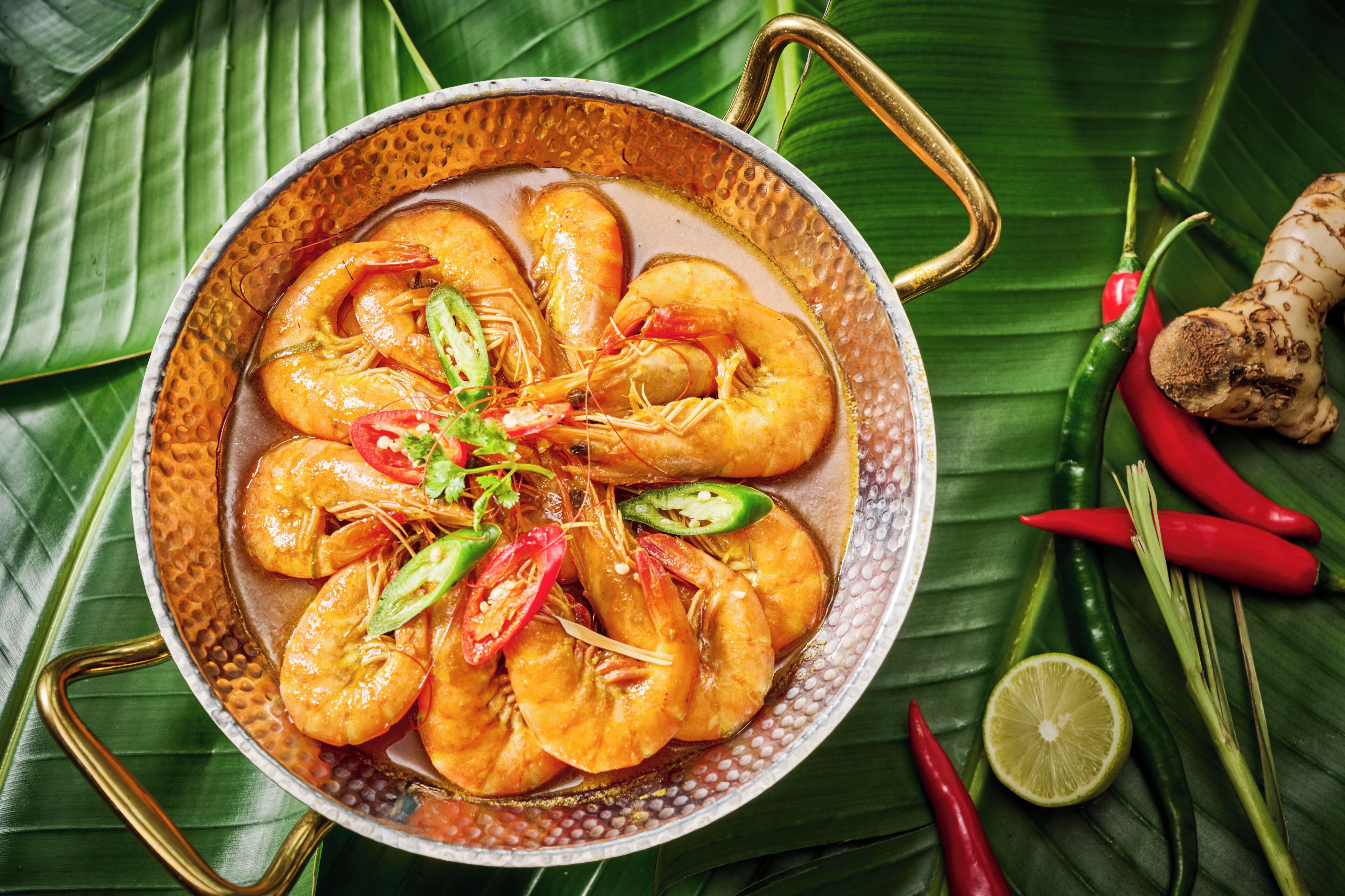 A pan of cooked shrimp garnished with chili peppers and herbs, placed on a banana leaf.