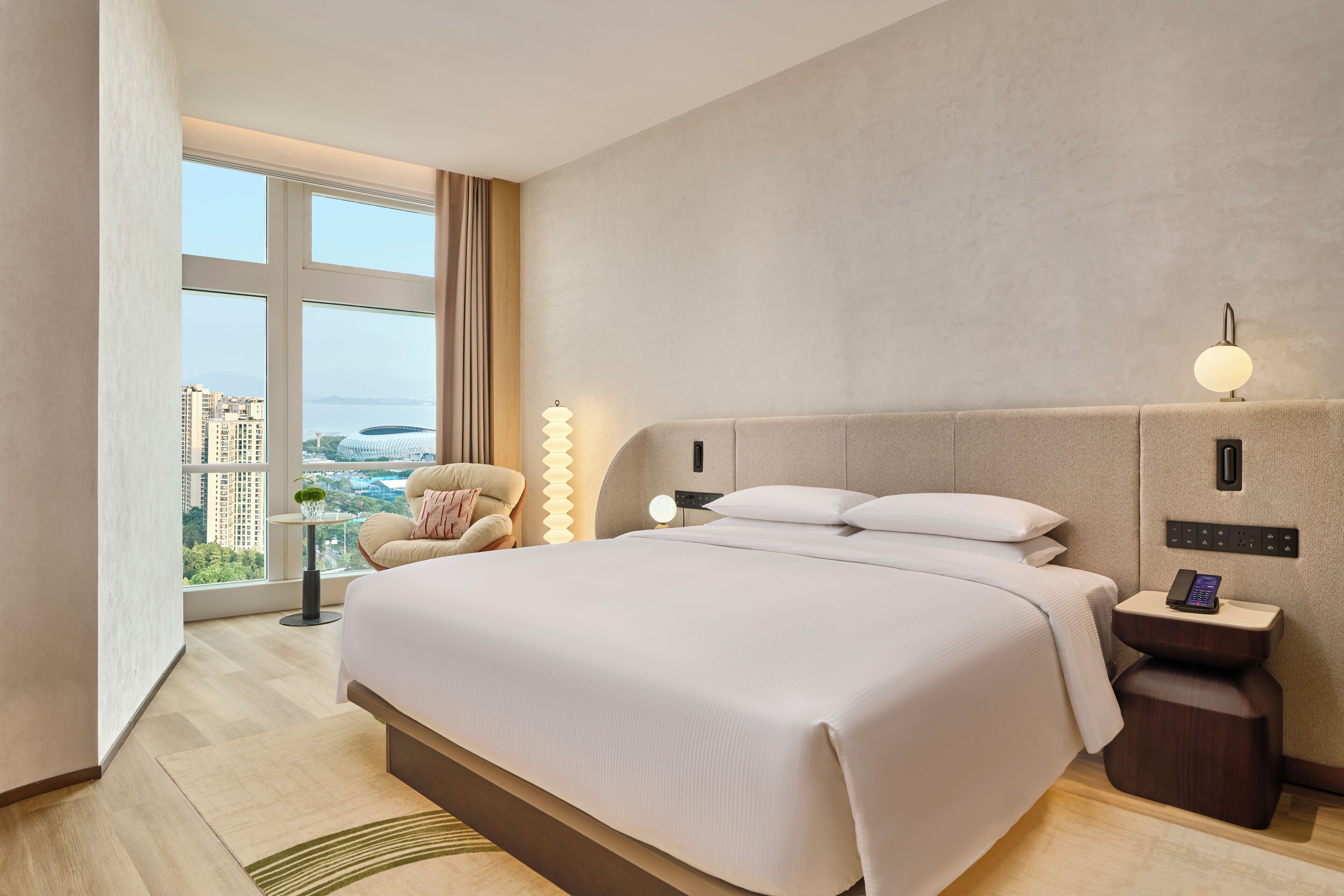 A modern hotel room with a large bed, a bedside table with a lamp, a phone, and a view of the cityscape.