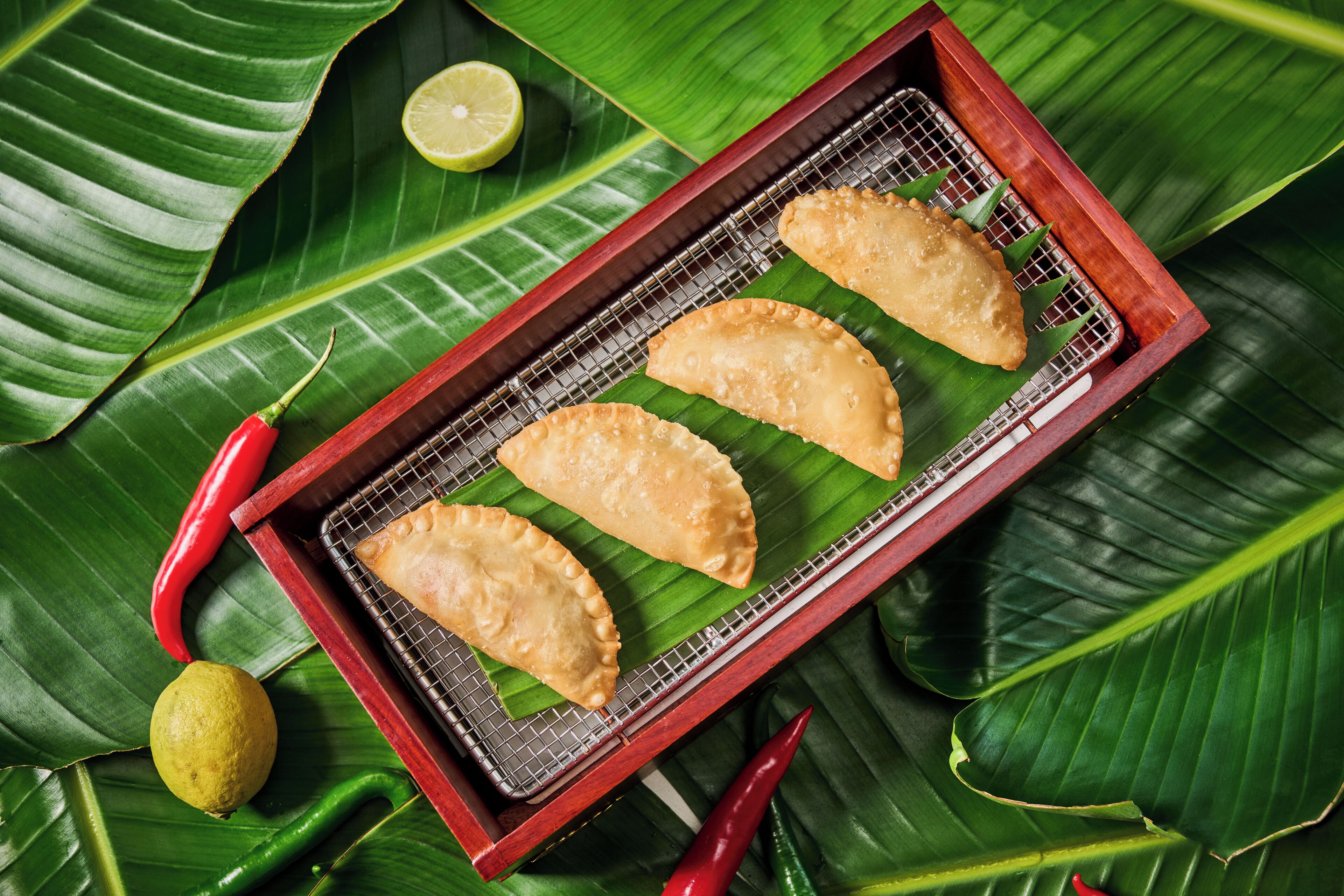Three fried pastries on a wire rack placed on a wooden tray, surrounded by green leaves and red chili peppers.