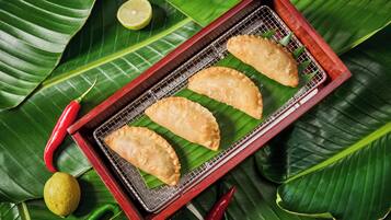 Three fried pastries on a wire rack placed on a wooden tray, surrounded by green leaves and red chili peppers.