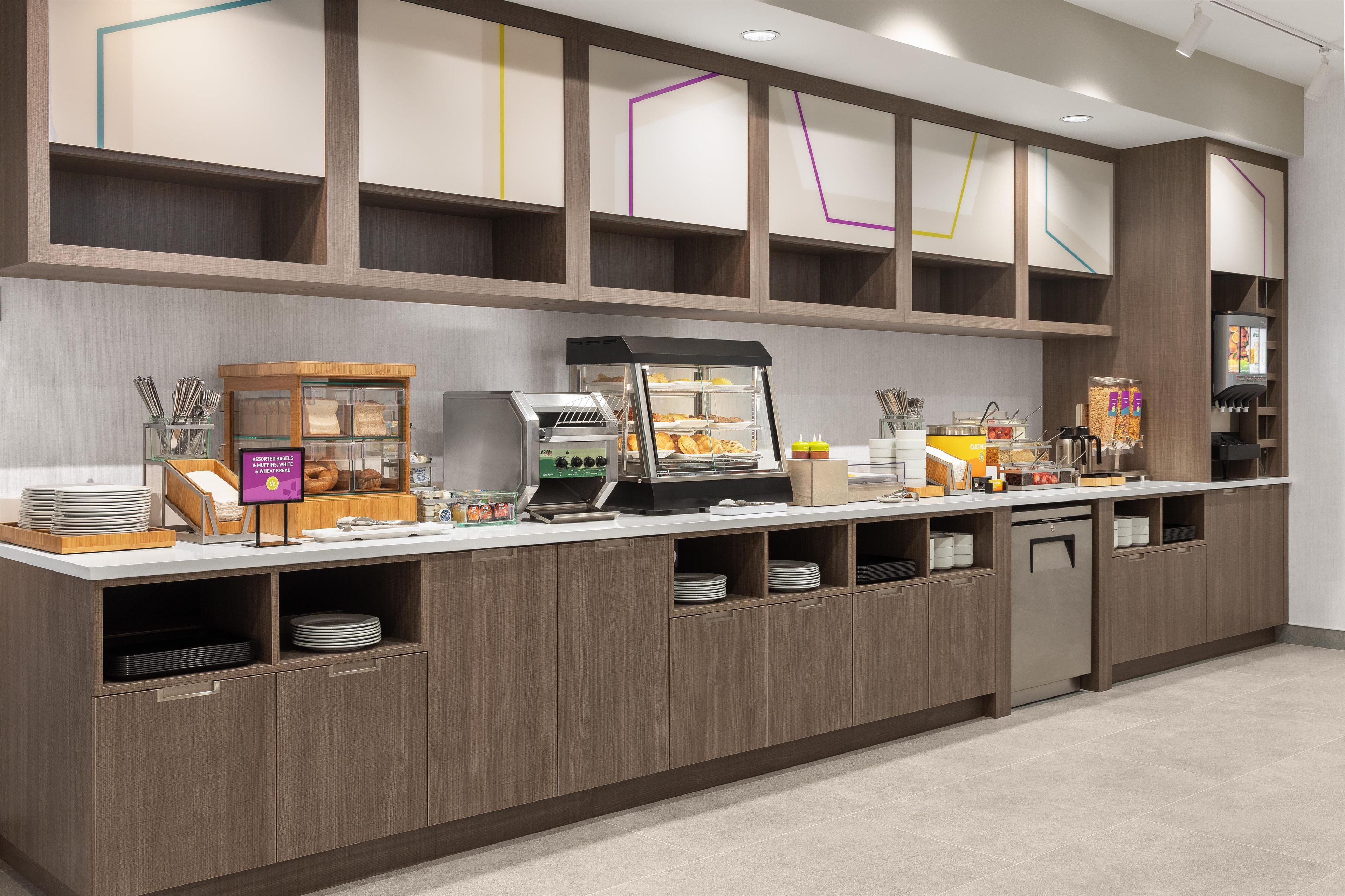 Estación de desayuno autoservicio con una máquina de café, un expositor de pasteles y un dispensador de bebidas.