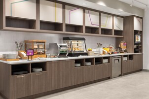 A self-service breakfast station with a coffee machine, a display case with pastries, and a beverage dispenser.