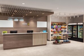 Zona de recepción de hotel con un mostrador de madera, dos sillas negras, un frutero y un letrero que dice "HOME". Hay estantes con aperitivos y un refrigerador con las etiquetas "FRÍO" y "MUY FRÍO".