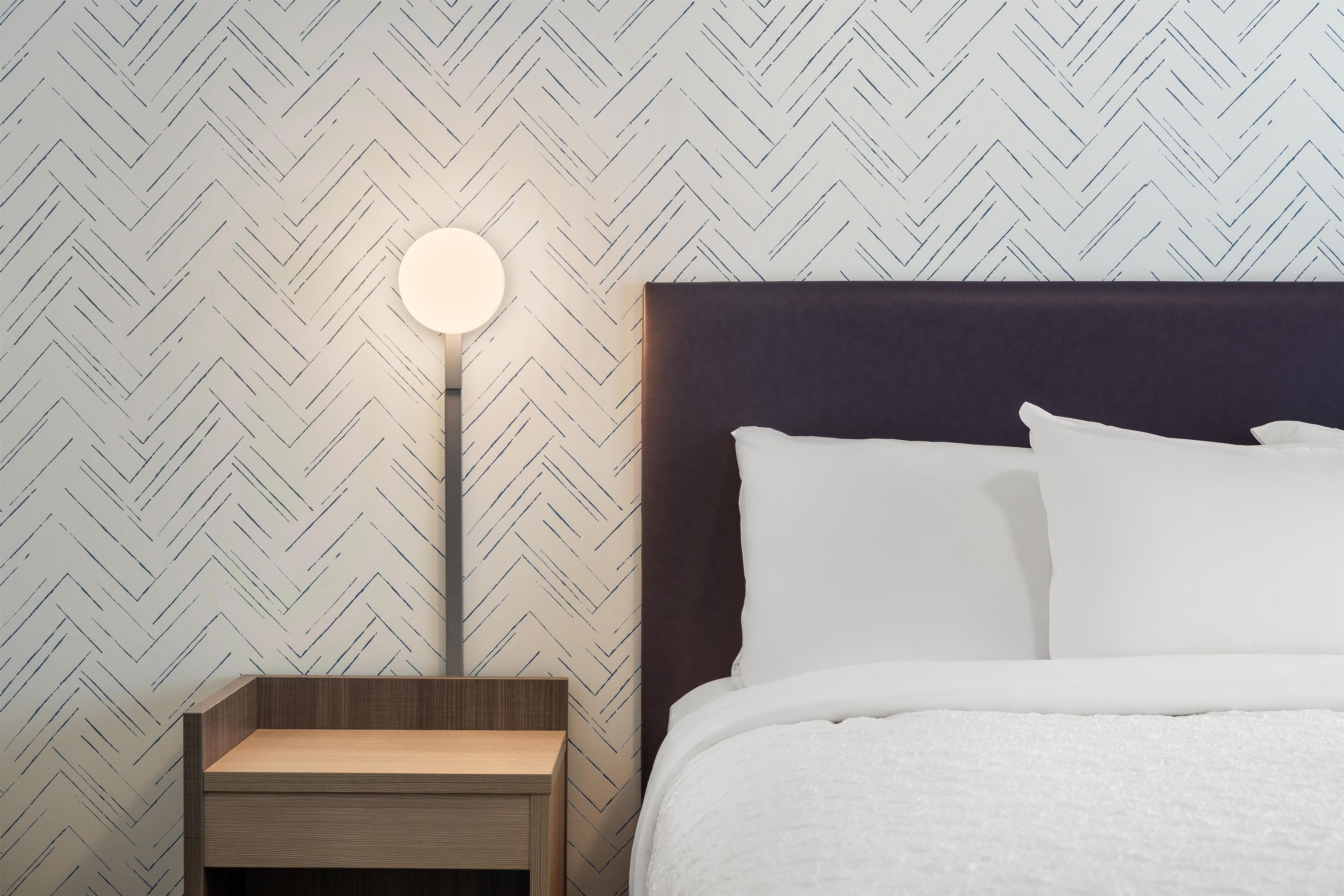 Una cama bien hecha con ropa blanca, una lámpara de noche redonda y una mesita de noche de madera.