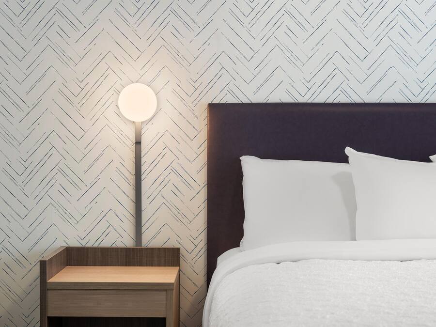 Una cama bien hecha con ropa blanca, una lámpara de noche redonda y una mesita de noche de madera.