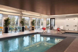Una piscina interior con paredes de vidrio transparente, suelo de baldosas y algunas sillas de descanso.