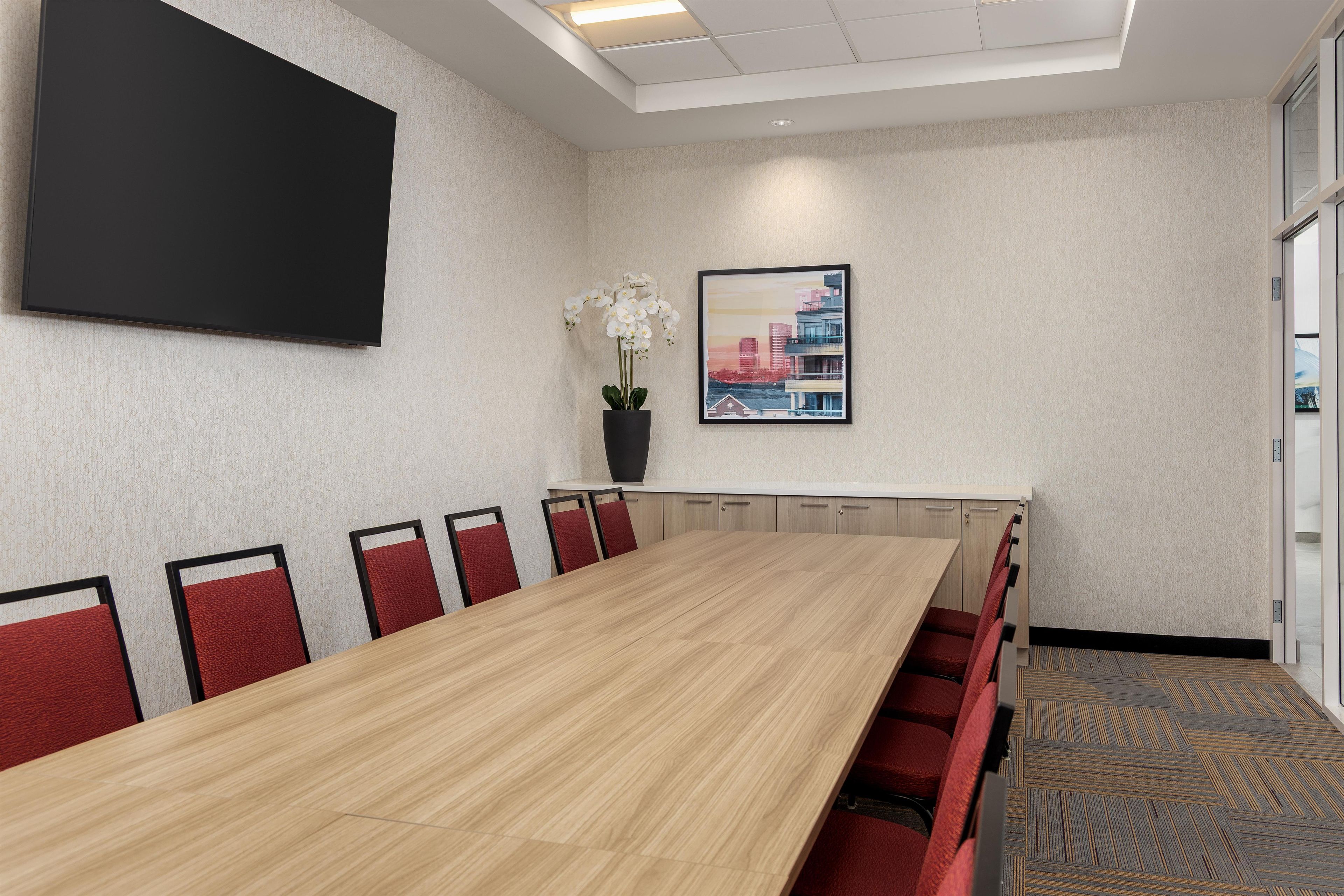 A conference room with a long table, red chairs, a potted plant, a framed cityscape picture, and a mounted screen on the wall.