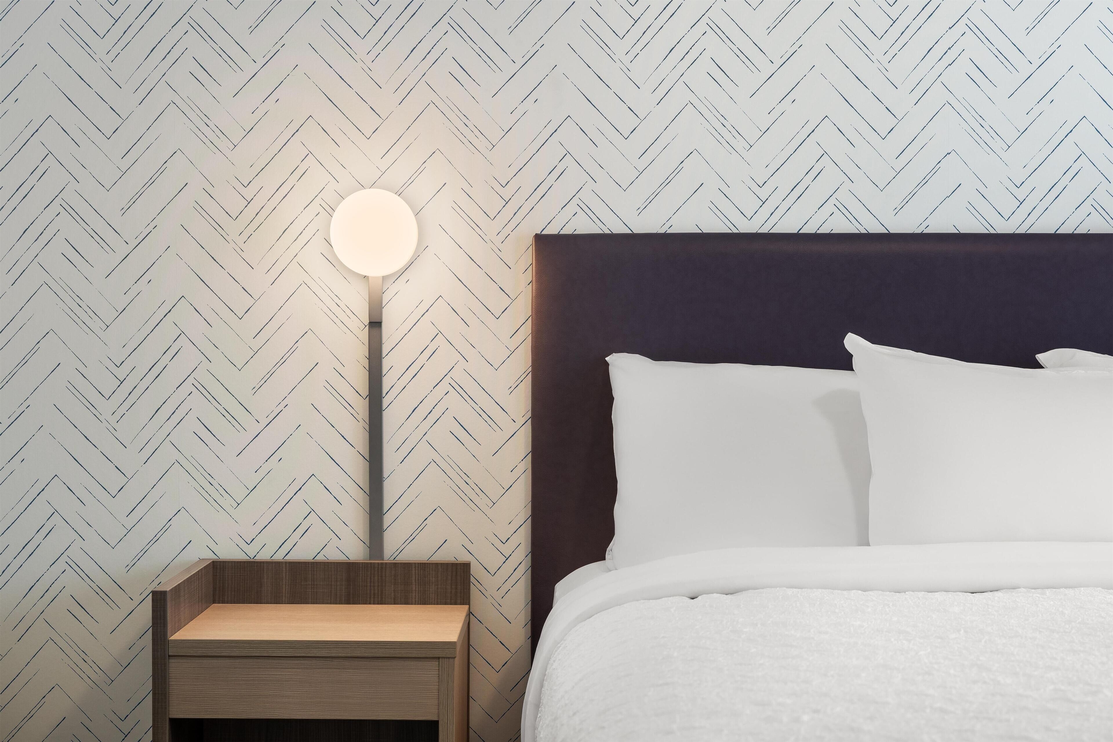 Un lit bien fait, avec du linge de lit blanc, une tête de lit sombre et une table de chevet sur laquelle se trouve une lampe ronde.