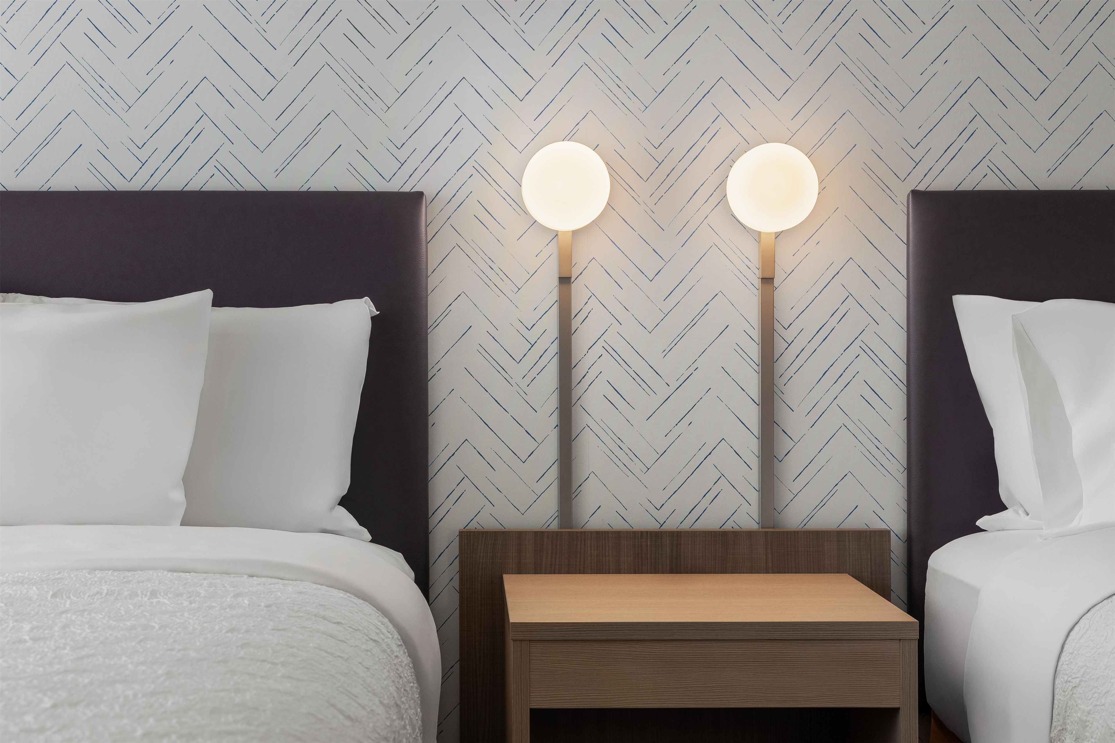 Habitación de hotel con dos camas, una mesita de noche y una pared con patrón de espiga.