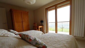 A bedroom with a large bed, a wooden wardrobe, a small table with a lamp, and a balcony with a view. - Gîte des Buis du Chardonnet (COGNY)