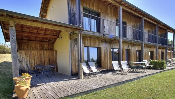 A two-story wooden building with a covered patio area, outdoor seating, and a view of the surrounding landscape. - Gîte des Buis du Chardonnet (COGNY)
