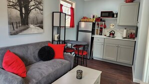 A compact living space with a grey sofa, red cushions, a small table, a kitchen area with white cabinets, and a framed tree artwork on the wall. - Charming 1-bedroom cottage -- That tiny house on Center Hill Lake. Pet friendly! (Baxter)