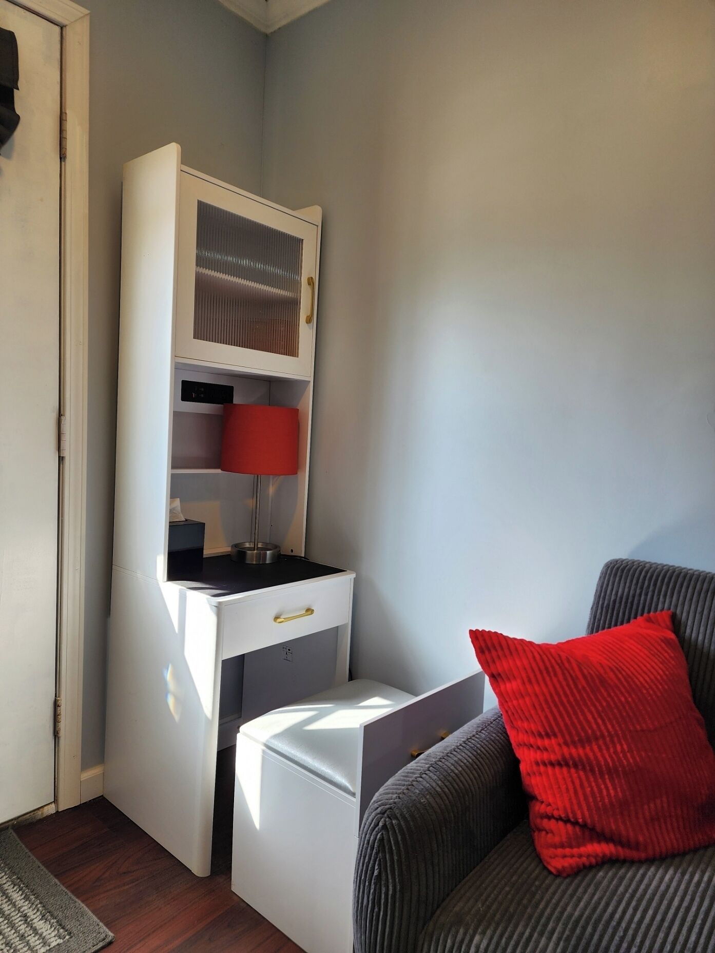 A compact room with a white cabinet, a red lamp, and a grey sofa with a red cushion.