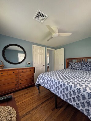 Une chambre avec une commode en bois, un miroir rond, un lit avec une literie à motifs, un ventilateur de plafond et une porte donnant sur une autre pièce.
