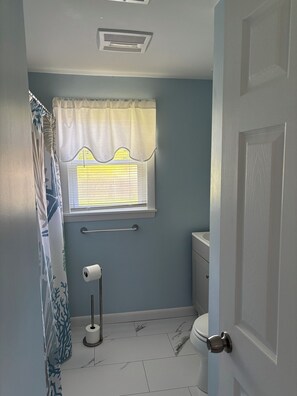 Une salle de bain comprenant des toilettes, une fenêtre avec un rideau, un porte-serviettes et un support à papier toilette.