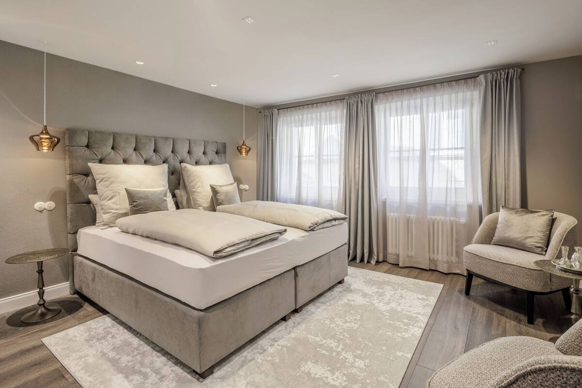 Un dormitorio moderno con una cama grande, una mesita de noche, una silla y una ventana con cortinas translúcidas.
