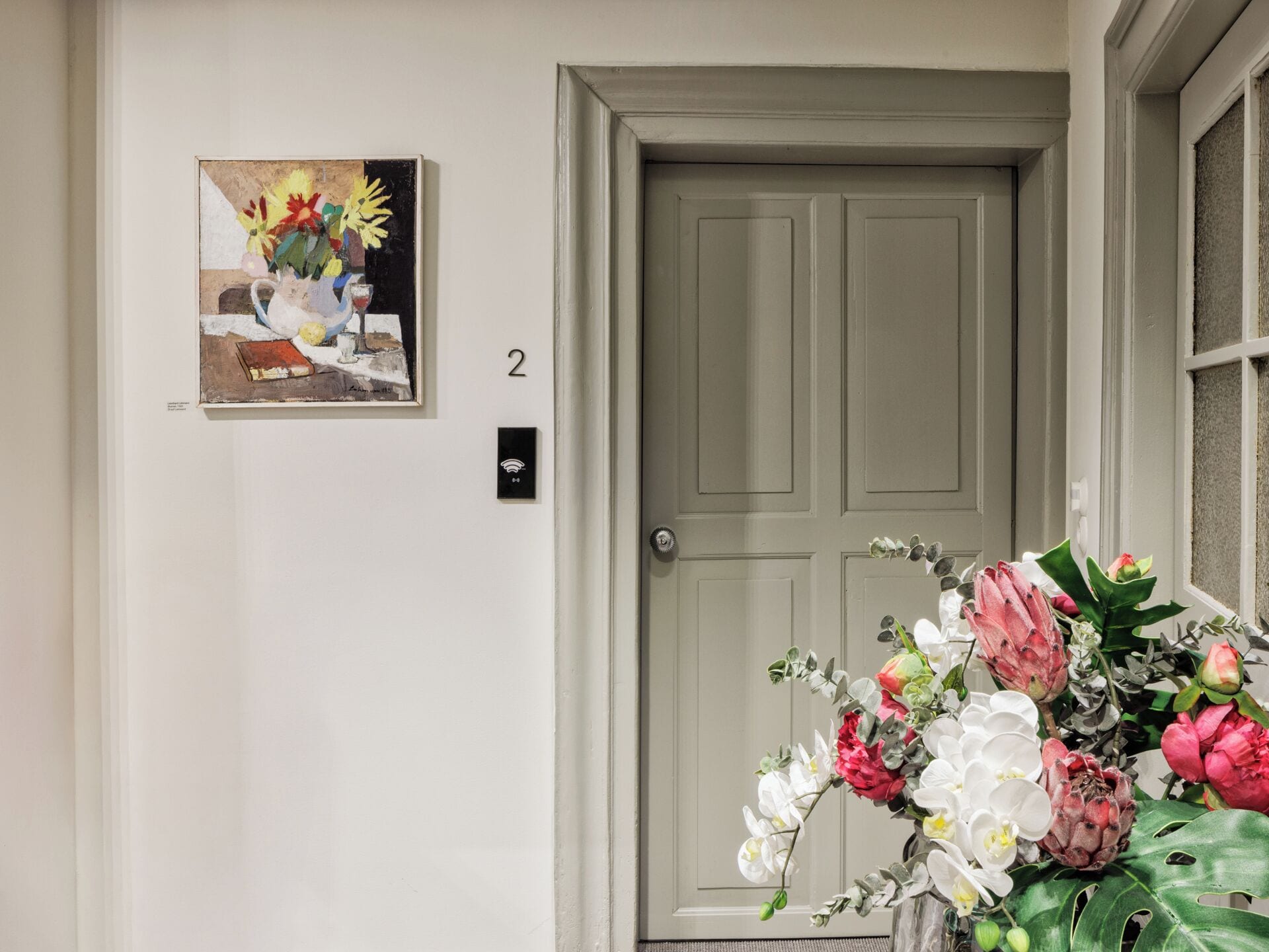 Una puerta con un cartel con el número 2, un cuadro de flores y un florero con flores.