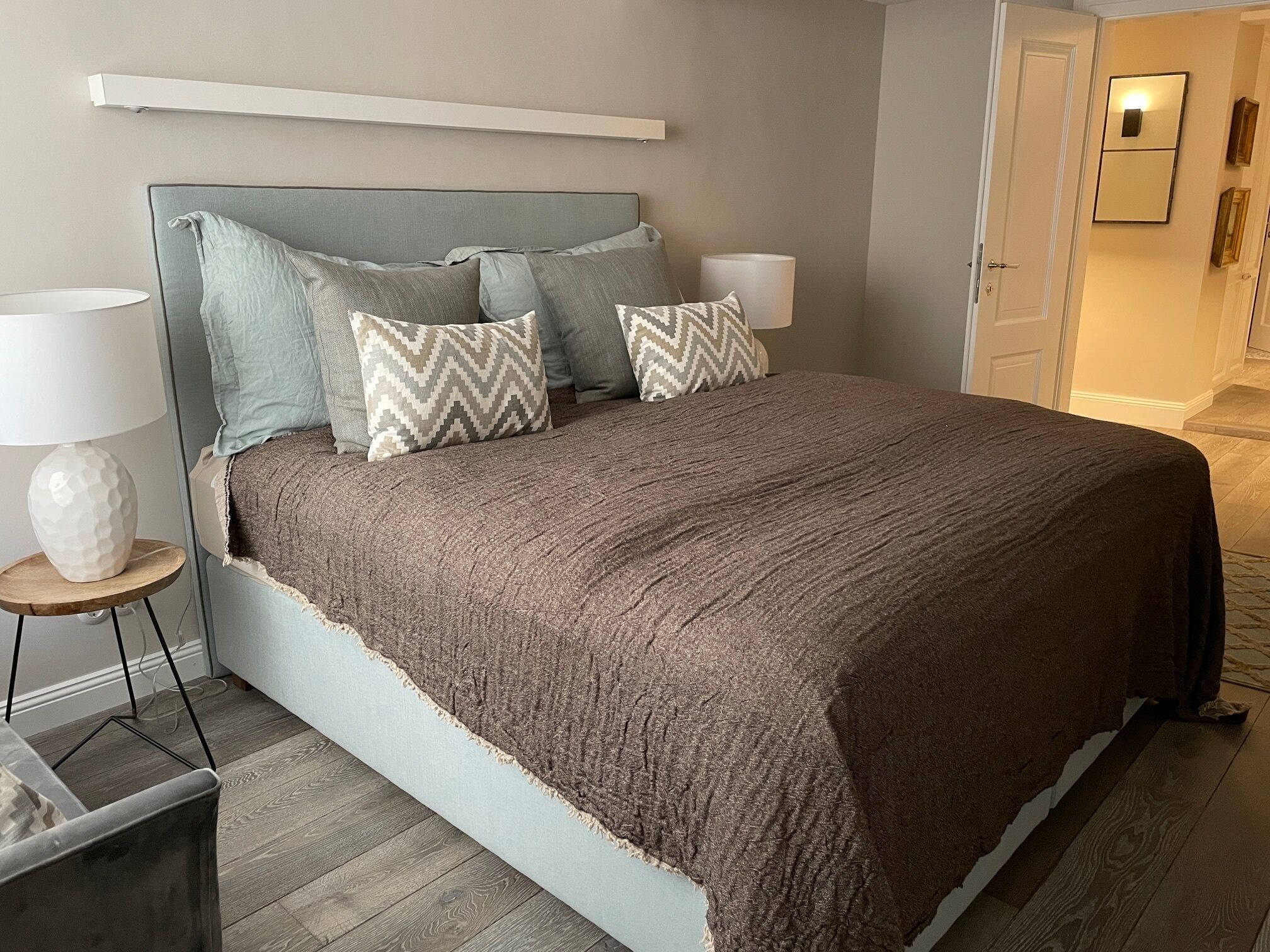 Un lit bien fait, avec une tête de lit grise, plusieurs oreillers et une couverture marron. On y trouve une table de chevet avec une lampe et un vase décoratif. La pièce est dotée d’un plancher en bois et d’une porte qui mène à un autre espace.