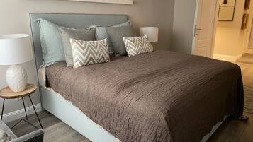 A neatly made bed with a grey headboard, multiple pillows, and a brown blanket. There is a bedside table with a lamp and a decorative vase. The room has wooden flooring and a door leading to another area.