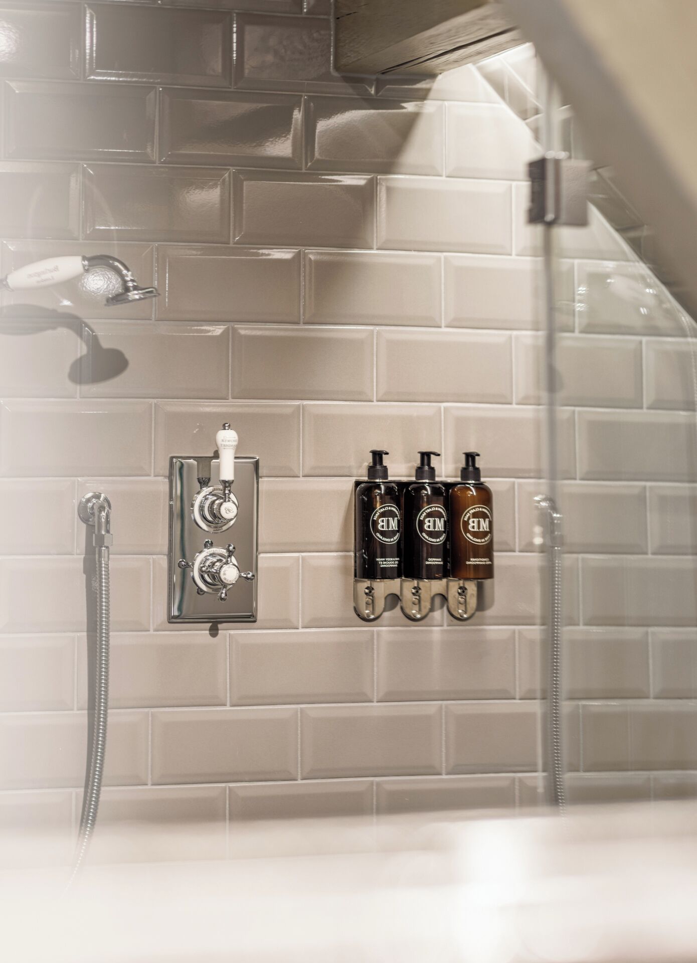 Zona de ducha con pared de azulejos, ducha de mano, grifo de pared y tres botellas de champú.