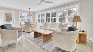 A spacious living room with a white sofa, two wicker chairs, a wooden coffee table, and a ceiling fan. Large windows provide natural light and a view of the outdoors.