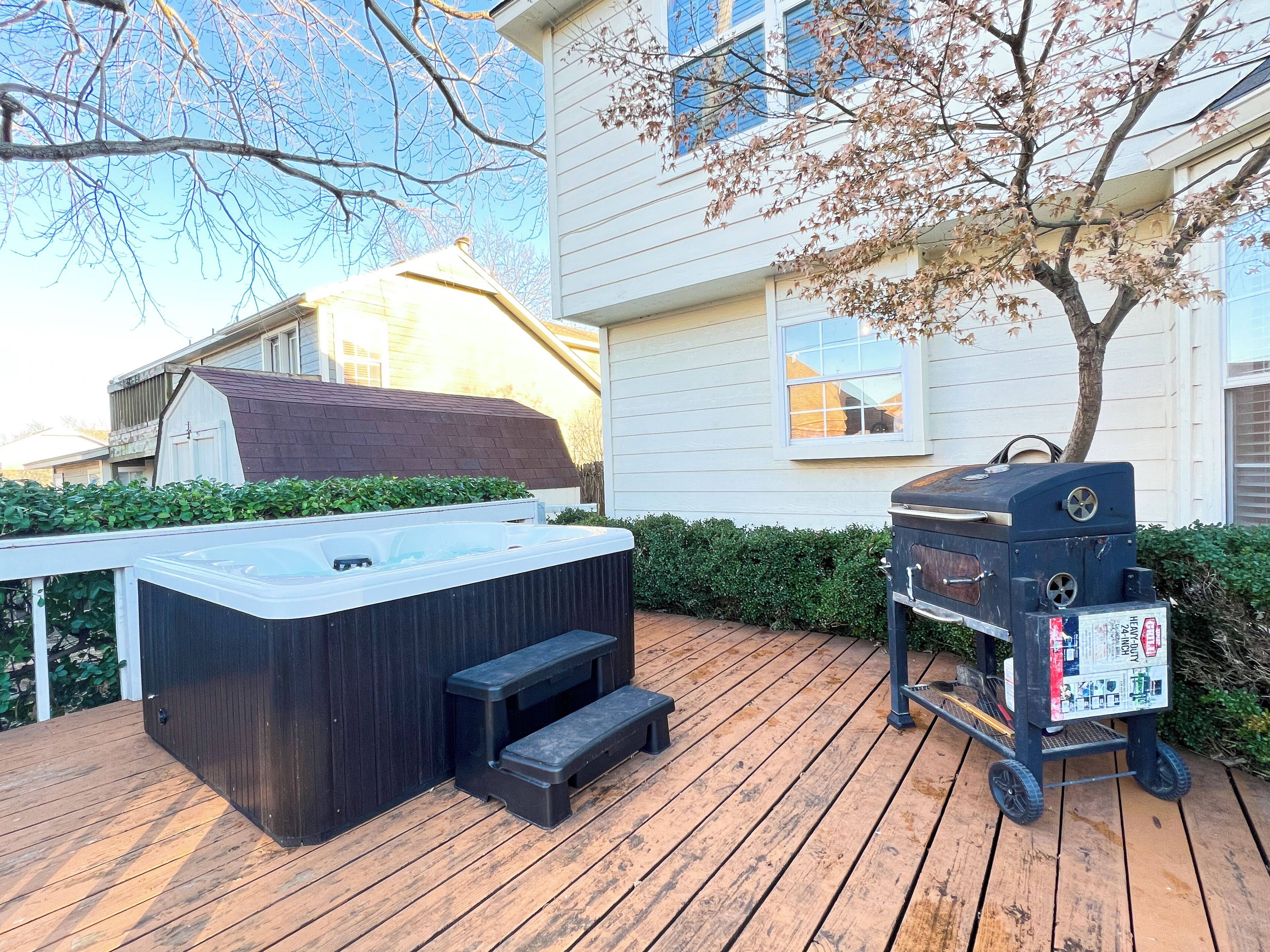 Un jacuzzi sur une terrasse en bois, accessible par des marches, un barbecue et une maison en arrière-plan.