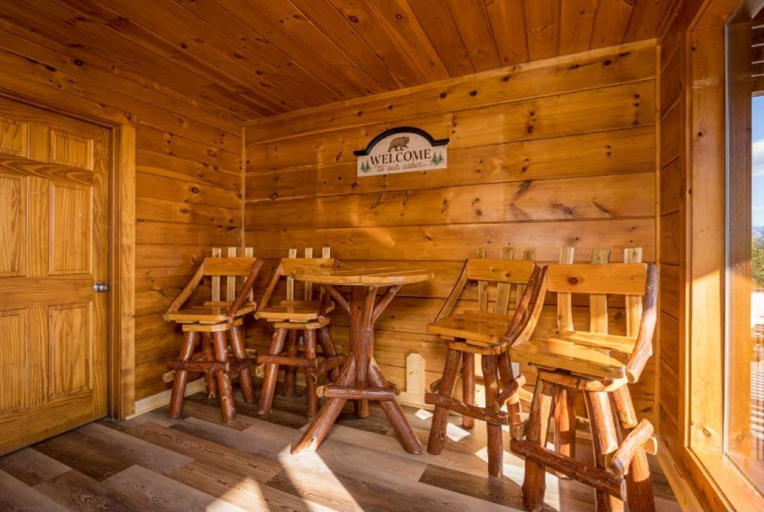 A wooden cabin interior with a 'Welcome' sign, wooden furniture, and a herringbone-patterned floor.