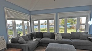 A large gray sectional sofa with a matching ottoman, a small wooden table, and a view of a lake and outdoor furniture through large windows.