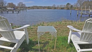 Two white Adirondack chairs facing a calm lake with docked boats and a grassy area in the foreground.
