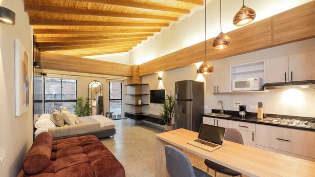 A modern kitchen with wooden beams, a dining area with a laptop, and a cozy seating area with a sofa and cushions. - Tranvia 40 (Medellín)