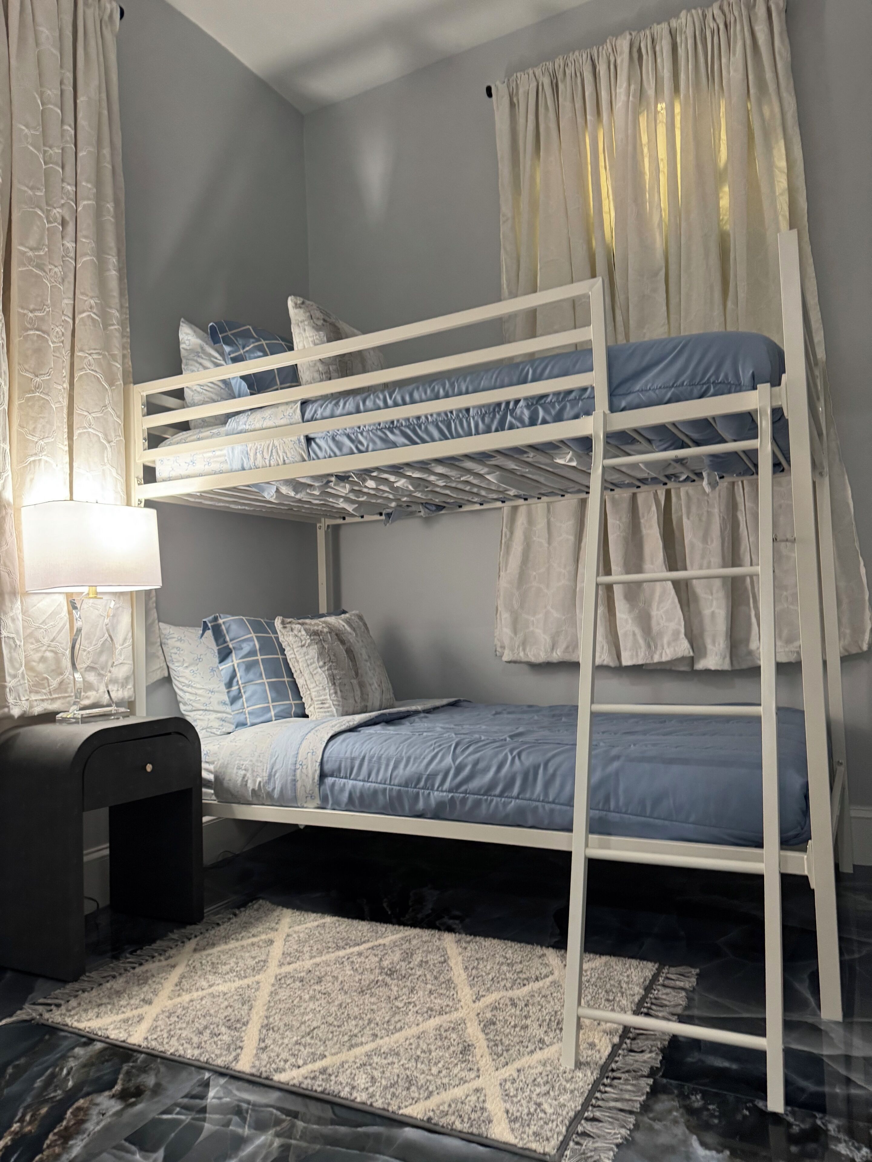 A bunk bed with a blue mattress, white bedding, and patterned pillows. There is a small bedside table with a lamp, a rug on the floor, and a window with curtains.
