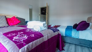 Une chambre d’hôtel avec deux lits, l’un recouvert d’une couvre-lit violette et l’autre d’une couvre-lit bleue. On aperçoit des serviettes sur les lits, et une porte est visible au fond.