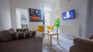 Un salon comprenant un canapé, une table à manger avec des chaises, un téléviseur fixé au mur et une fenêtre avec des rideaux.