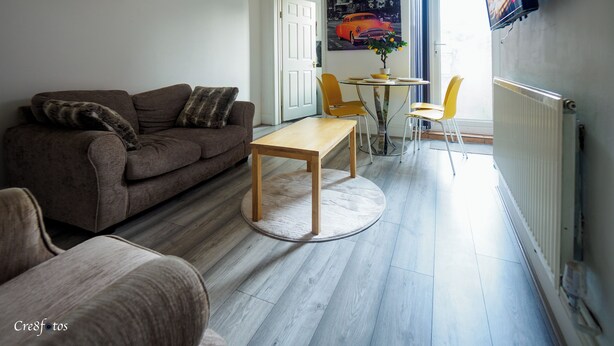 Un salon avec un canapé marron, une table basse en bois et des chaises jaunes. On y trouve une porte qui mène à une autre pièce et un radiateur fixé au mur.