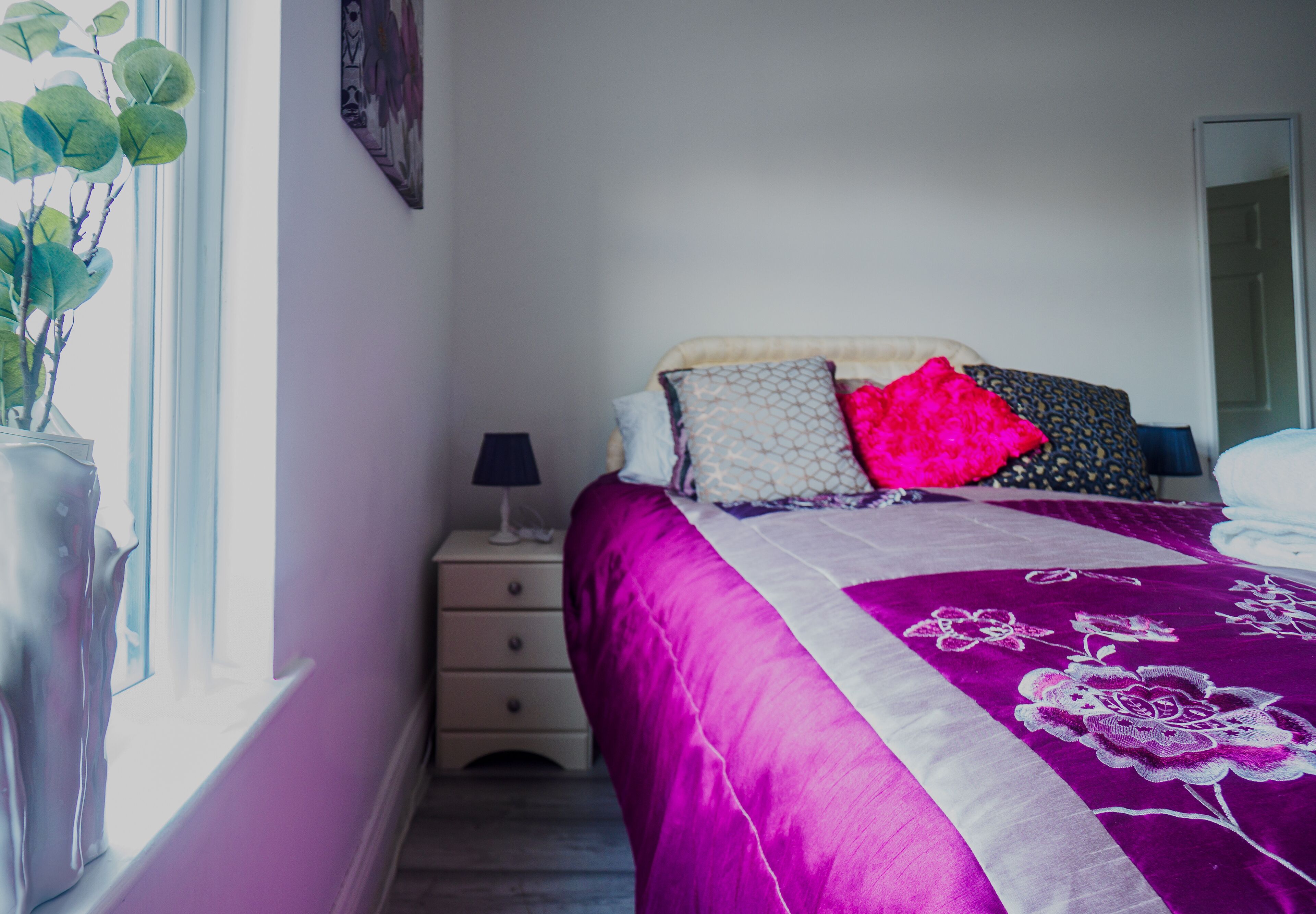 A bedroom with a bed, a nightstand, a lamp, and a window with a plant.