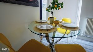 Un coin repas avec une table en verre, des chaises jaunes, une plante en pot et un tableau représentant une voiture accroché au mur.