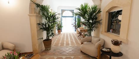 A spacious hallway with a checkered floor, two large potted plants, a beige armchair, a small round table, and a decorative vase.