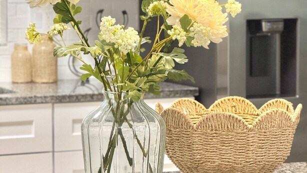 A kitchen with a clear glass vase containing flowers and a woven basket on the counter.